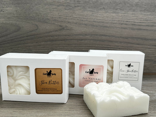 3 various bars of natural soap with a white and gold packaging, labeled 'Shea Butter, All Natural and Coco Shea Butter', along with its box and a small container variant, all displayed on a wooden surface.