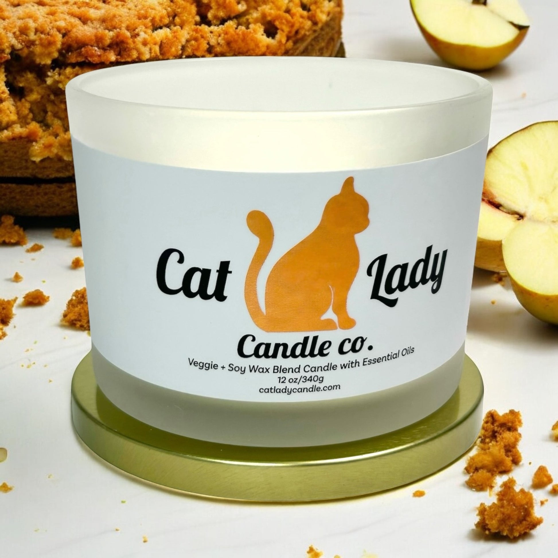 Candle with 'Cat Lady Candle Co.' label surrounded by an apple strudel cake, cinnamon and sugar crumbles, and apple slices on a light background