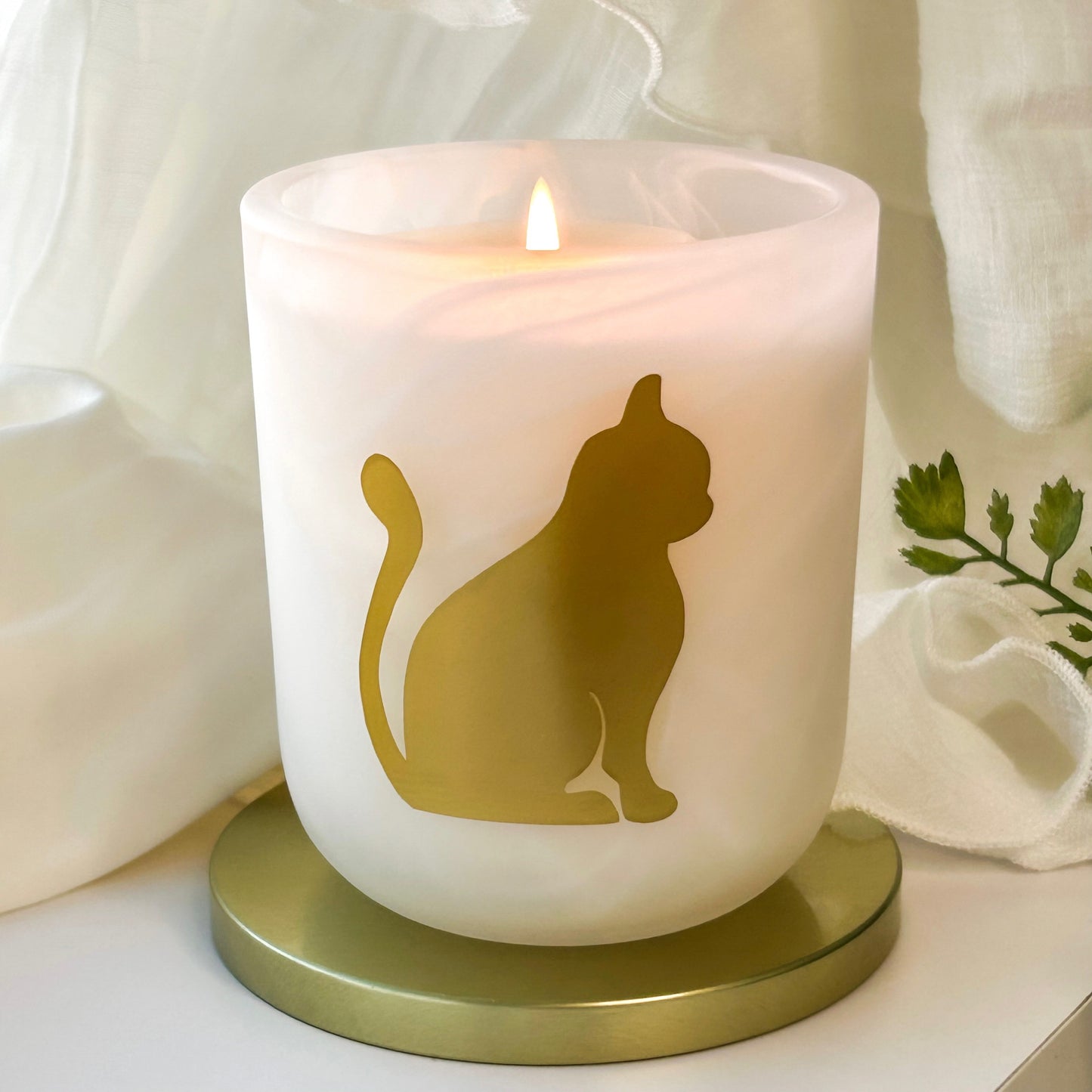Southern Comforts sweet potato pie scented candle with cinnamon and freshly baked sweet potato pie.
A frosted white candle in a cylindrical glass holder with a gold cat silhouette design, lit with a small flame and set on a round gold base against a soft white draped fabric background.