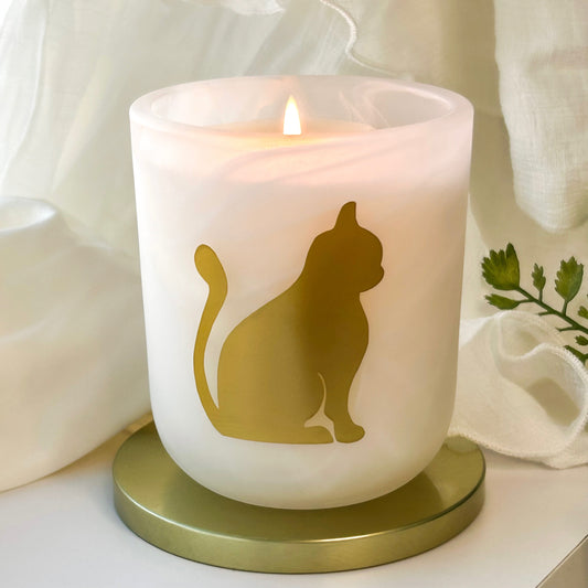 Homestead Hearth pine cedar musk scented candle by Cat Lady Candle Co. A frosted white candle in a cylindrical glass holder with a gold cat silhouette design, lit with a small flame and set on a round gold base against a soft white draped fabric background.