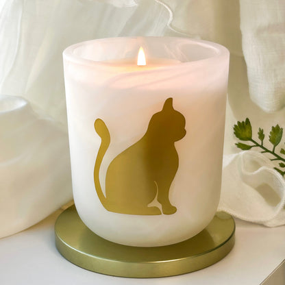 Velvet Ambrosia orange vanilla gourmand scented candle by Cat Lady Candle Co. A frosted white candle in a cylindrical glass holder with a gold cat silhouette design, lit with a small flame and set on a round gold base against a soft white draped fabric background.